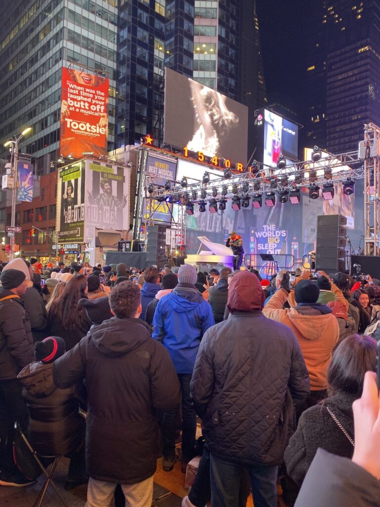 Crowd gathered at night in Times Square for the World's Big Sleep Out event, with a performer on a lit stage